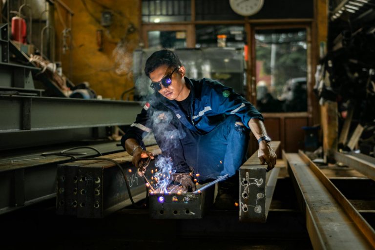 A dedicated welder wearing safety gear operates a welding torch in a bustling industrial workshop.