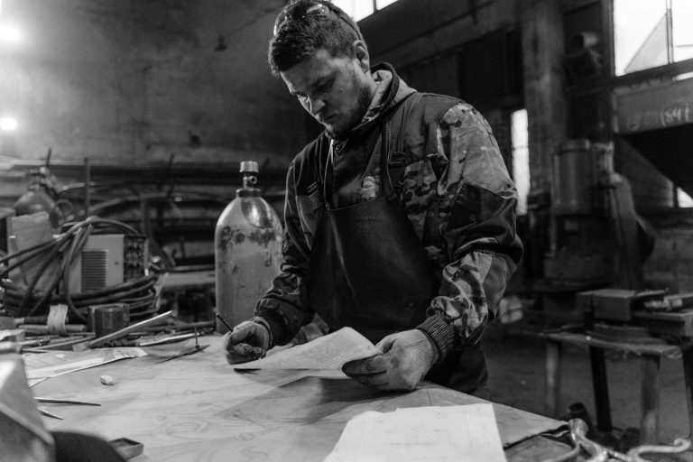 A focused blacksmith in a workshop reviews design plans, surrounded by tools and metal.
